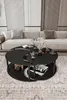 A black, Yin Yang-shaped coffee table is shown in a modern living room setting with a light-colored sofa and decorative panels on the wall. The table has two tiers and holds a vase of flowers, books, and cups. Produktbild-Vorschau