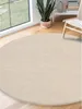 A round, cream-colored rug is placed on a wooden floor in a room with a large window. A potted plant and a chair are visible in the background. Produktbild-Vorschau