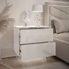 A white, high-gloss nightstand with two drawers is shown next to a bed. The nightstand has chrome legs and is decorated with a bust and a lamp. Produktbild-Vorschau
