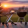 A barbecue grill and a folding table are set up on a patio during sunset. Produktbild-Vorschau