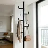 A black telescopic coat rack with several hooks, holding a jacket, two necklaces, and a handbag. The coat rack is positioned in a hallway or entryway, with a glimpse of a living room and a large window in the background. Produktbild-Vorschau