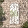 A decorative mirror with an ornate metal frame is mounted on a rustic stone wall, with greenery visible in the reflection and to the sides. Voir les images du produit