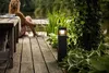 A person sits on a wooden deck next to a lake, with a modern black garden light illuminating the plants. Anteprima dell’immagine prodotto