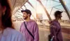 A man wearing in-ear headphones and a hoodie stands in a modern, glass-enclosed walkway. Produktbild-Vorschau