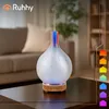 An aroma diffuser with LED lights is placed on a coffee table in a living room setting. The background shows a couch and bookshelves. Produktbild-Vorschau