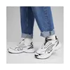 A person wearing jeans and white socks is standing in a pair of white Puma sneakers. Voir les images du produit