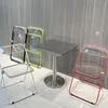 A small square table with four transparent chairs around it, set against a grey, wood-paneled wall. Anteprima dell’immagine prodotto