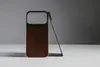 A brown iPhone case with a black frame is standing upright, leaning against a thin metal stand. The background is a gradient of light gray to dark gray. Produktbild-Vorschau