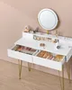 A white vanity table with gold legs, featuring a round illuminated mirror and organized drawers filled with makeup. Produktbild-Vorschau