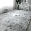 A fluffy, light gray rug is laid out on a wooden floor, with a white polka-dot cushion and a book placed on it. A plant and a white shelf unit are visible in the background. Podgląd zdjęcia produktu