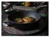 A frying pan with food in it, placed on a wooden surface, with bowls of dip and a stack of food in the background. Produktbild-Vorschau