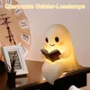 A cute ghost-shaped LED lamp is reading a book, placed on a table next to a framed picture and a clock. Produktbild-Vorschau