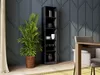 A tall, narrow black bookshelf filled with various decorative items and glassware, placed next to a large potted plant in a woven basket. A dining table and chairs are partially visible on the right. Produktbild-Vorschau