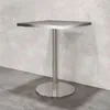 A square-top table with a single pedestal base and a round foot, set against a textured wall and concrete floor. Anteprima dell’immagine prodotto