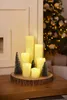 A Christmas-themed centerpiece with LED candles and miniature trees on a wooden base, with a decorated Christmas tree in the background. Produktbild-Vorschau