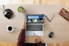 A person's hands are using a laptop on a wooden desk, with a cup of coffee, a plant, a lamp, glasses, and pens nearby. Voir les images du produit