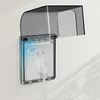 A close-up shot of a transparent electrical outlet cover with a plug inserted, mounted on a white wall. Produktbild-Vorschau