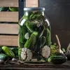 A large glass jar filled with pickles, garlic, and herbs, placed on a wooden surface with more pickles and garlic around it. A wooden crate is visible in the background. Podgląd zdjęcia produktu