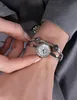 A close-up shot of a woman's wrist wearing a silver Guess watch with a white dial and black accents. The watch is part of a bracelet with decorative spheres. Produktbild-Vorschau