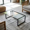 A glass coffee table with a black metal frame is placed on a rug in a living room. Sunlight streams in from a large window, casting shadows. Produktbild-Vorschau