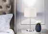 A bedside table with a lamp and a vase of flowers next to a bed. Náhled obrázku produktu