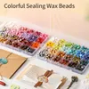 A collection of colorful sealing wax beads in compartmentalized boxes, along with envelopes and a wax seal stamp. Náhled obrázku produktu