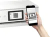 A hand holds a smartphone displaying a QR code, positioned next to a printer with a similar QR code on its screen. Podgląd zdjęcia produktu