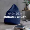 A navy blue beanbag chair is placed in a modern living room setting, with books and a small cup on the floor nearby. A side table with a vase and plant is visible in the background. Náhled obrázku produktu