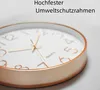A close-up of a wall clock with a rose gold frame and a white face, with German text overlayed. Produktbild-Vorschau