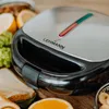 A close-up of a sandwich maker on a wooden surface with some food items around it. Anteprima dell’immagine prodotto