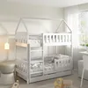 A white wooden bunk bed with a house-shaped frame, set up in a child's bedroom with soft lighting and a window in the background. Produktbild-Vorschau