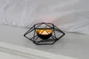 A black geometric candle holder with a lit tea light inside, placed on a white surface with a white fabric background. Podgląd zdjęcia produktu