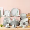 A collection of white ceramic dinnerware with green grass-like accents, arranged on a wooden surface against a pink background. Náhled obrázku produktu