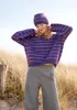 A woman wearing a purple striped sweater and hat stands in a field of tall dry grass. Produktbild-Vorschau