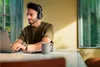 A man wearing headphones is working on a laptop at a table, with a mug and a phone nearby. Sunlight streams in from a window. Voir les images du produit