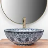 A decorative vessel sink with a blue and white floral pattern sits on a wooden surface, with a gold faucet and a round mirror in the background. Náhled obrázku produktu