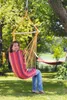 A woman is sitting in a colorful hammock chair hanging from a tree in a garden. Produktbild-Vorschau