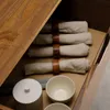 A close-up view of a drawer in a wooden sideboard, showing rolled napkins with wooden napkin rings, a white lidded container, and two small bowls. Produktbild-Vorschau