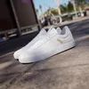 A pair of white Adidas sneakers are placed on a paved surface with a blurred background of palm trees and a bicycle. Produktbild-Vorschau