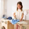 A young girl is playing with a wooden toy washing machine and ironing board, pretending to do laundry. Produktbild-Vorschau