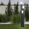 A modern outdoor lighting fixture with three columns of varying heights, illuminated at the top, set in a garden with manicured grass, ornamental grasses, and a white wall in the background. Produktbild-Vorschau