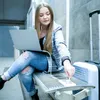 A young woman is sitting on a bench in a public space, using a laptop and charging her phone. Podgląd zdjęcia produktu