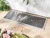 A metal grate cover for a basement window well, partially covered with leaves, set into a patio with flowers and grass nearby. Produktbild-Vorschau