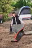 A man is setting up a sleeping bag next to a tent in a wooded area. Podgląd zdjęcia produktu
