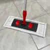 A mop with a white microfiber pad and red and black handle is placed on a tiled floor. Produktbild-Vorschau