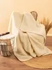 A cream-colored wool blanket is draped over a chair, with a small side table holding magazines next to it. A woven rug is on the floor. Produktbild-Vorschau