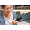 A woman is smiling while looking at her smartphone, sitting on a couch with pillows. Produktbild-Vorschau