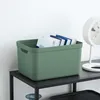 A green storage box filled with books sits on a black shelf next to a white fan. Produktbild-Vorschau