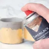A hand is spraying a can of KREUL Zaponlack onto a gold and gray pot. Produktbild-Vorschau