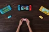 A top-down view of hands holding a pink gamepad, surrounded by Nintendo Switch consoles and controllers on a wooden table. Náhľad obrázku produktu